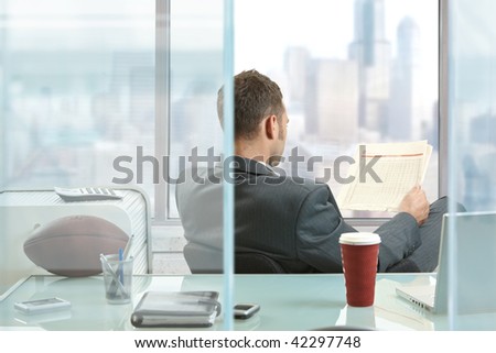 Relaxed businessman  sitting at desk in front of office windows, reading nwespaper.