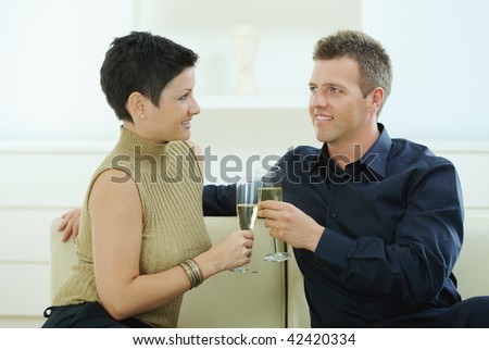 Love couple clinking champagne glasses at home on sofa. Smiling and looking at each other.