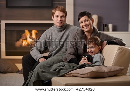 Happy family sitting on sofa at home in front of fireplace, looking at camera, smiling.