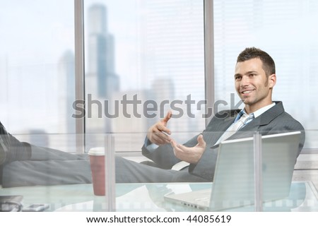 Relaxed businessman  sitting at desk in front of office windows, talking to somebody.