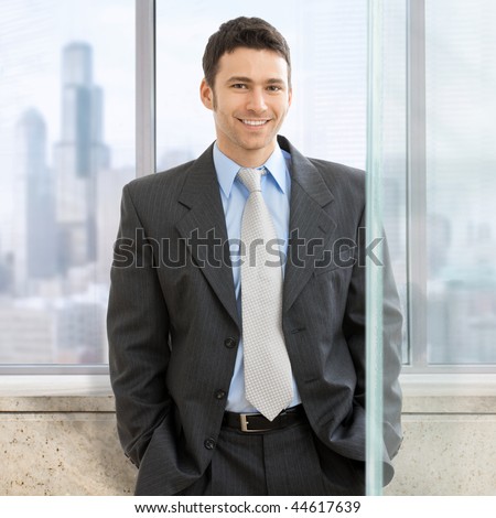 Portrait of businessman standing with hands in pocket in front of office windows, smiling.