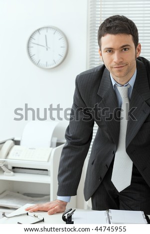 Young businessman wearing grey suit, standing behind office desk, smiling.