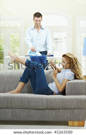Relaxed young woman sitting on couch filing her nails, man ironing in the background. Selective focus on woman.