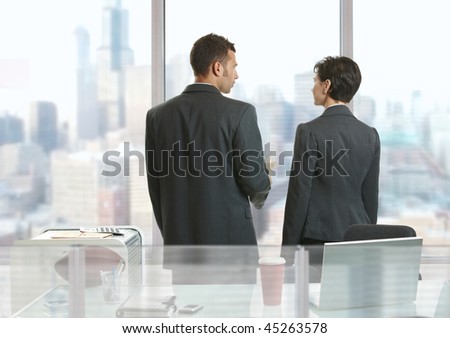Two businesspeople standing at desk in office, looking out the windows and talking.