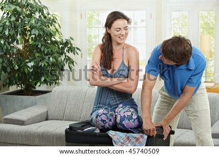 Happy couple packing for holiday, woman trying to close suitcase full with clothes.