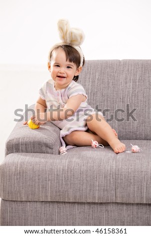 Baby girl in easter bunny costume, sitting on grey couch playing with toy chicken and easter eggs, laughing. Isolated on white background.
