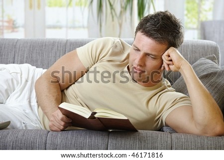 Handsome man in causal wear smiling lying on sofa reading handheld book.