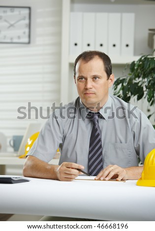 Engineer working at office - planning on blueprint on desk, looking away, thinking.