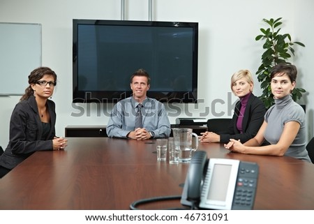 Business people sitting around meeting table in board room, waiting, looking at camera. Blank space on screen for your logo or text.