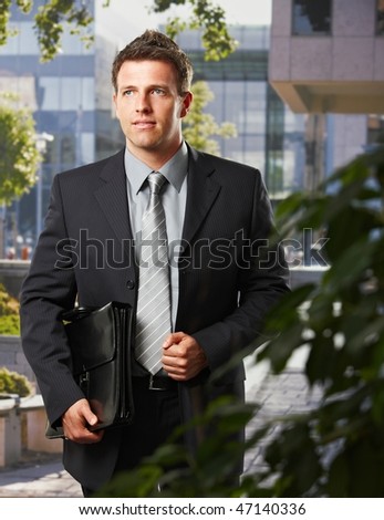 Smart executive in suit standing on office building courtyard holding briefcase.