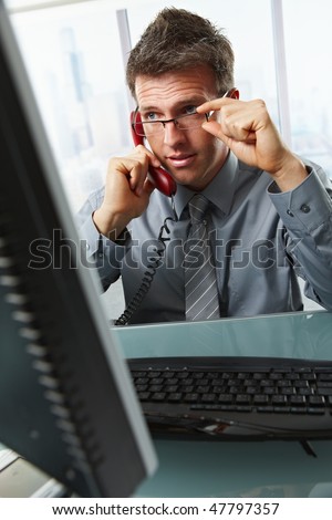 Determined businessman discussing computer work on landline phone while looking at screen at office desk.