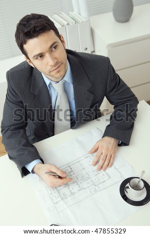 Architect wearing grey suit sitting at office desk, writing notes on floor plan, looking up. Overhead shot.