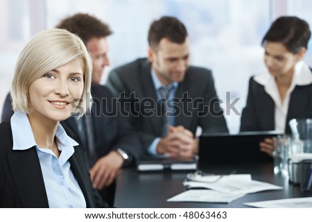 Portrait of mid-adult businesswoman smiling at camera with colleagues at meeting in background,