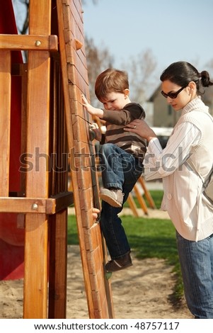 Mother helping little boy to climb wooden climbing wall on playground, sunlit outdoor.
