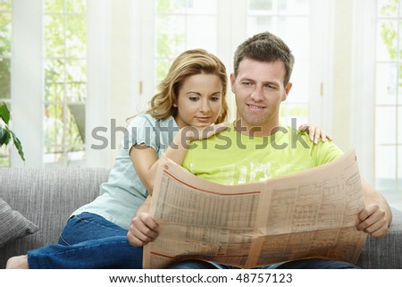 Love couple reading newspaper together on couch at home, smiling.
