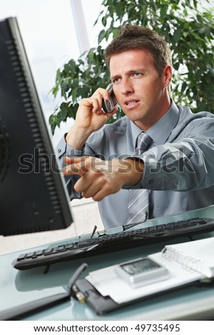Troubled businessman pointing at screen disussing work on mobile phone sitting at office desk.