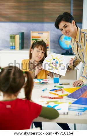 Teacher and elementary age schoolgirl showing colorful paining to classmate in art class in primary school classroom.
