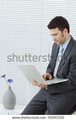 Businessman sitting on office cabinet holding using laptop compute in hand, working.