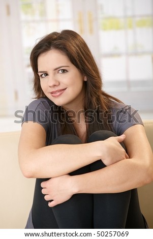 Portrait of smiling girl sitting on sofa with arms around legs pulled up, looking at camera.
