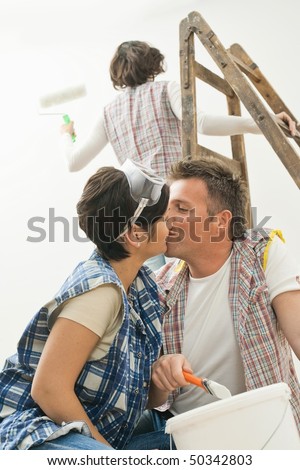 Couple kissing at home renovation, holding paint brush, female friend standing on ladder painting wall with paint roller.