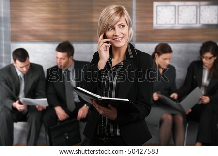 Happy young businesswoman standing in office hallway, holding personal organizer, talking on mobile phone.