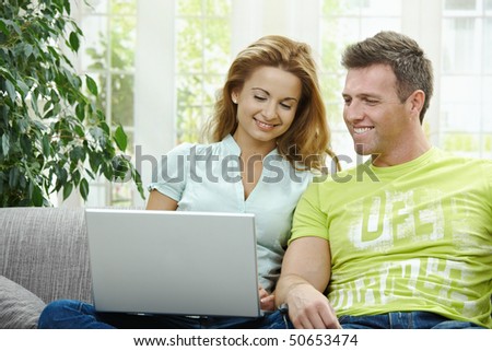 Couple using laptop computer at home together, looking at screen, smiling.