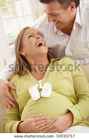 Happy expecting parents laughing, pregnant woman sitting on sofa with baby shoes on belly, man embracing looking down.