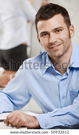 Happy businessman sitting at table in meeting room, looking at camera, smiling.