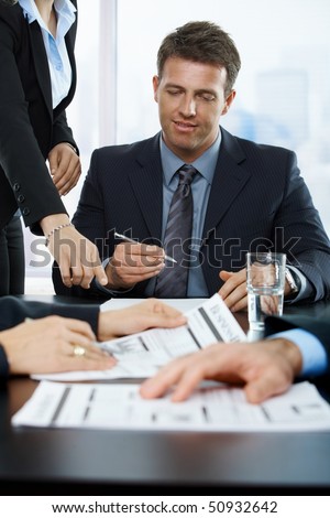 Smiling executive signing contract in office, assistant pointing at paper,