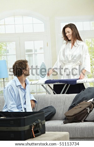 Young businessman packing for business trip. Woman ironing in the background.