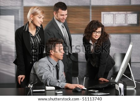 Businessman sitting at office desk, looking at screen together with colleagues, smiling.
