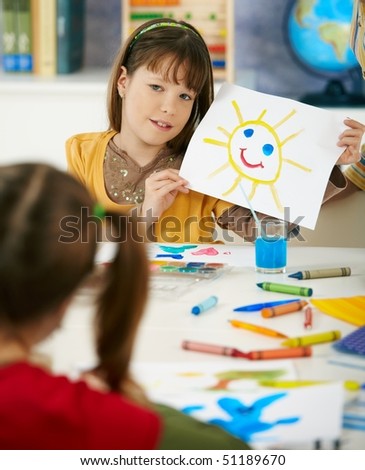 Portrait of elementary age schoolgirl showing colorful paining to classmate in art class in primary school classroom.