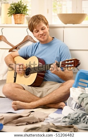 Lazy young guy playing guitar sitting on floor instead of doing housework.