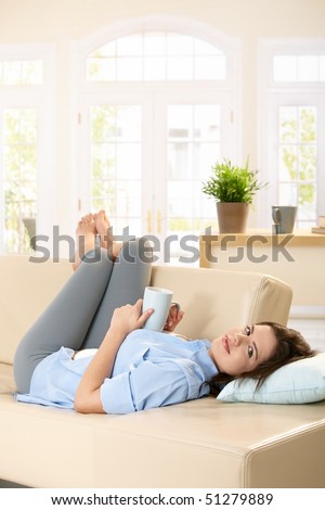 Attractive woman relaxing on couch with feet up, looking at camera, holding tea cup, smiling.