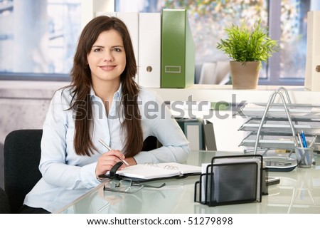 Smiling girl working in office sitting at desk with organizer, looking at camera.