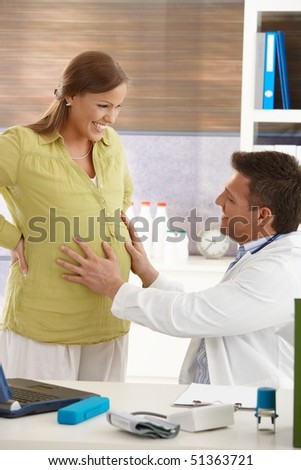 Smiling doctor touching belly of happy pregnant woman at examination in consulting room.