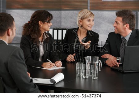 Young businesswoman explaining business problem, sitting at meeting table in office, others looking at her.