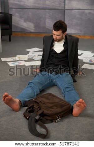 Trendy office worker sitting on office floor bare feet, surrounded with papers, documents and  laptop bag.