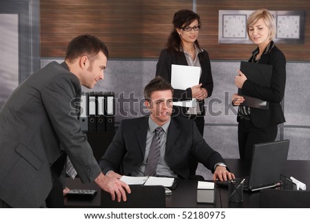 Businesspeople working at desk, boss sitting at desk, colleagues looking at laptop computer screen.