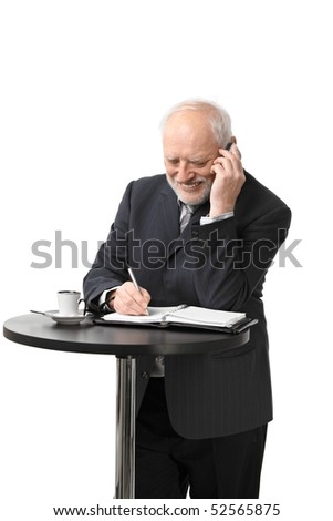 Portrait of happy senior businessman standing at coffee table talking on mobile phone, isolated on white.