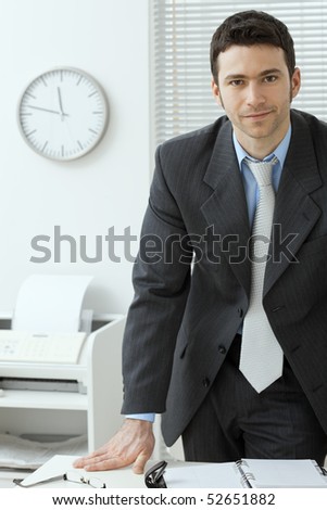 Young businessman wearing grey suit, standing behind office desk, smiling.