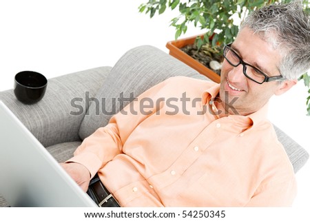 Happy man wearing orange shirt sitting on couch, browsing internet on laptop computer, smiling.