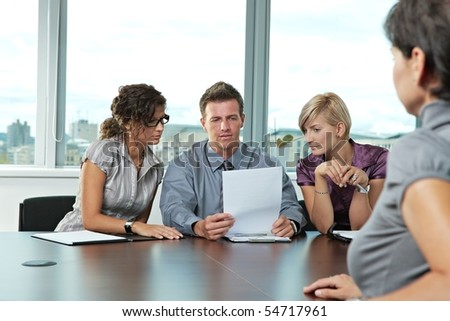 Panel of business people sitting at table in meeting room conducting job interview looking at documents.