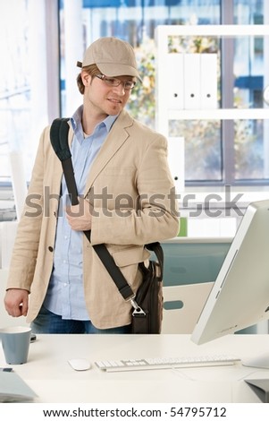 Young businessman wearing laptop bag leaving office, looking back to screen on desk.