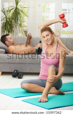 Young woman doing dumbbell exercise sitting on fittness mat at home, smiling. Man reading newspaper in background.