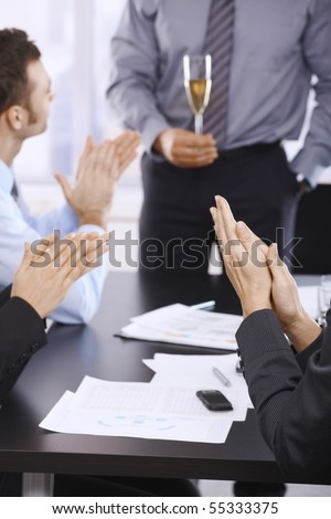 Businesspeople celebrating success with champagne in office, clapping hands. Focus on hand in front.?