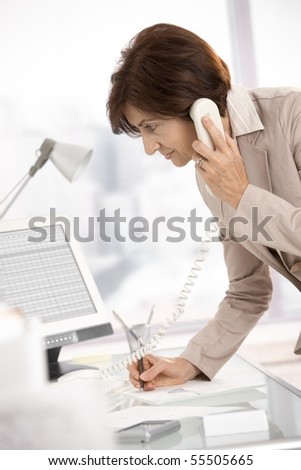 Senior businesswoman working in office, standing at desk, talking on landline phone, taking notes.