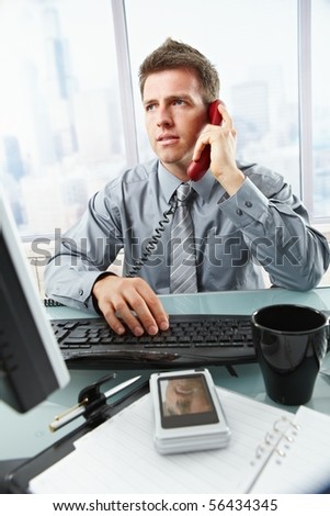 Determined businessman discussing computer work on landline phone while looking at screen typing on keyboard at office desk.