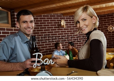 Portrait of young couple in bar, drinking beer, smiling at camera, friends in background.