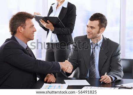 Businessmen shaking hands at meeting, sitting at table, assistant in background holding organiser.